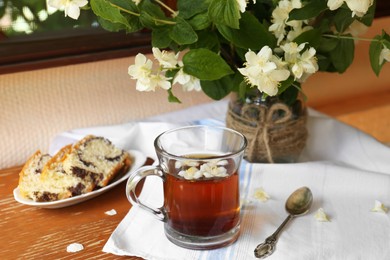 Photo of Glass cup of aromatic tea, tasty dessert and beautiful jasmine flowers on wooden table