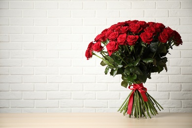 Luxury bouquet of fresh red roses on wooden table near white brick wall, space for text