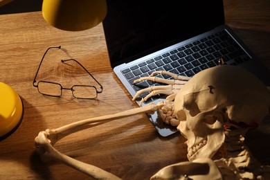 Photo of Waiting concept. Human skeleton sleeping at wooden table with laptop