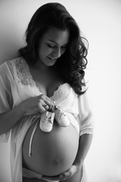 Young pregnant woman in lace nightgown holding baby shoes on light background, black and white effect