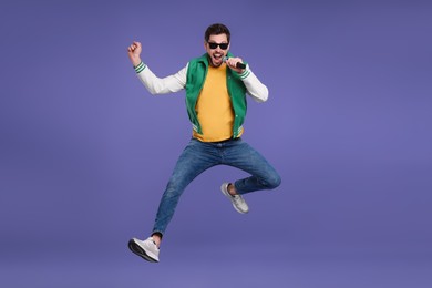 Photo of Handsome man wearing sunglasses with microphone singing and jumping on purple background