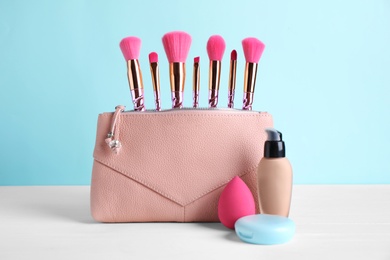 Photo of Bag with makeup brushes and cosmetic products on white table against light blue background