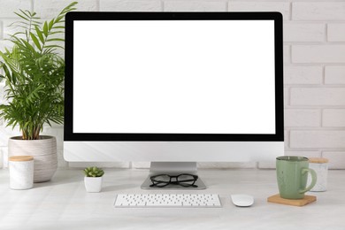 Photo of Office workplace with computer, cup, glasses, stationery and houseplant on light table near white brick wall