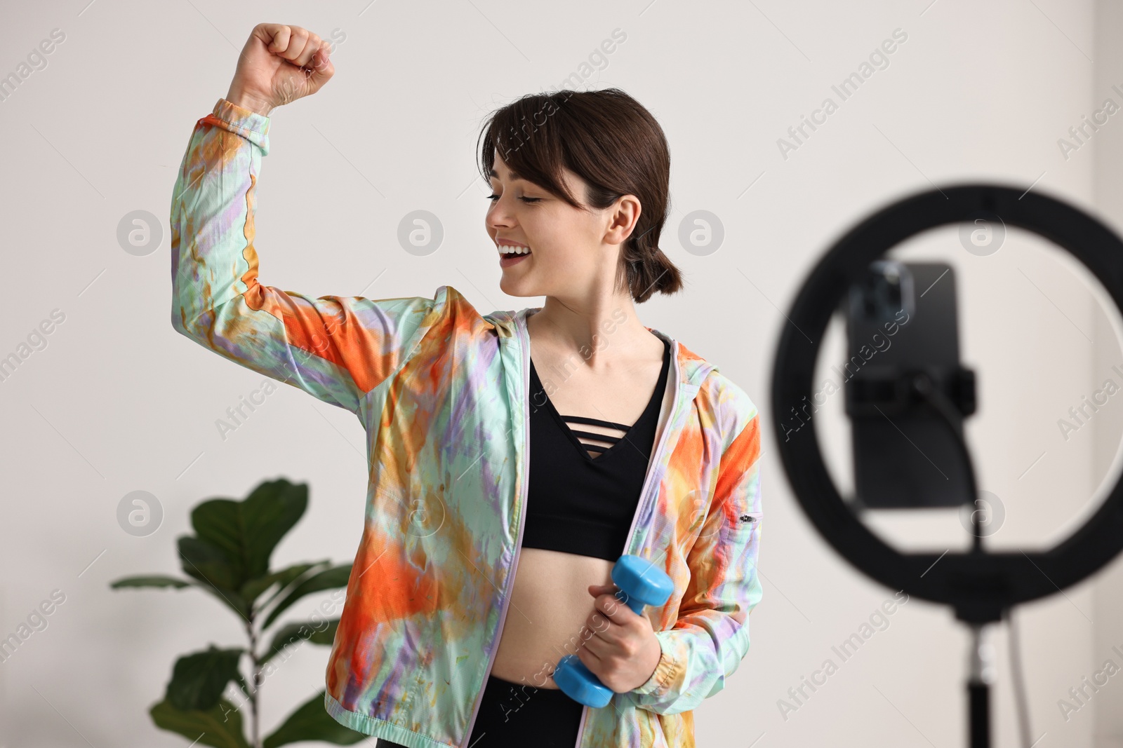 Photo of Happy sports blogger holding dumbbell while streaming online fitness lesson with smartphone at home