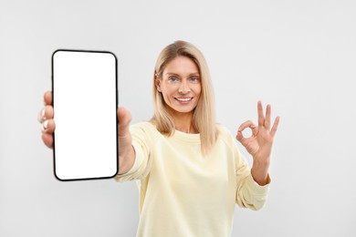 Happy woman holding smartphone with blank screen and showing OK gesture on white background