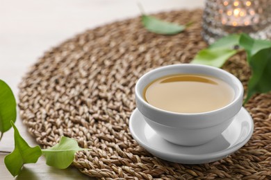 Photo of Green tea in white cup with leaves and wicker mat on table, closeup