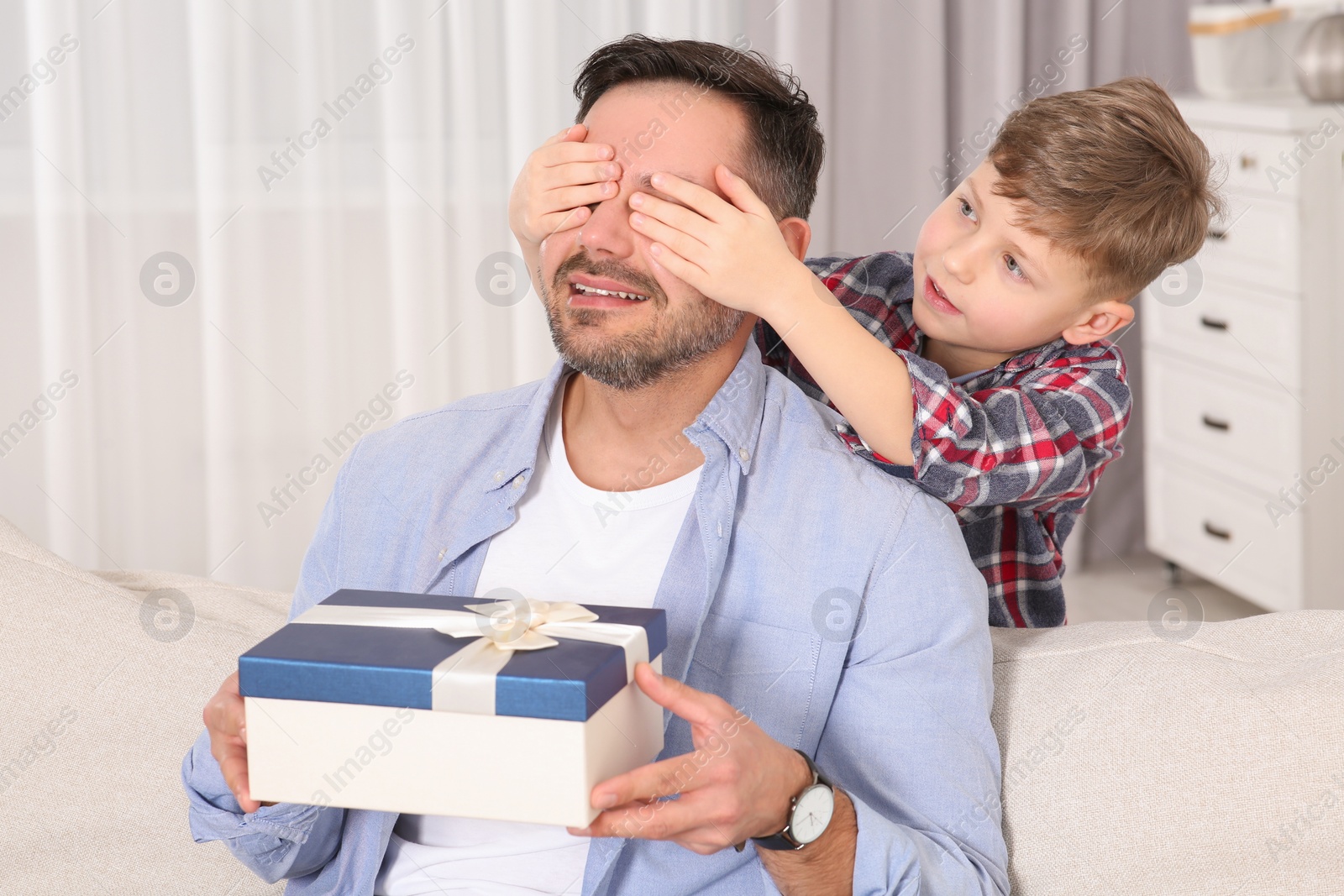 Photo of Cute little boy presenting his father with gift on sofa at home