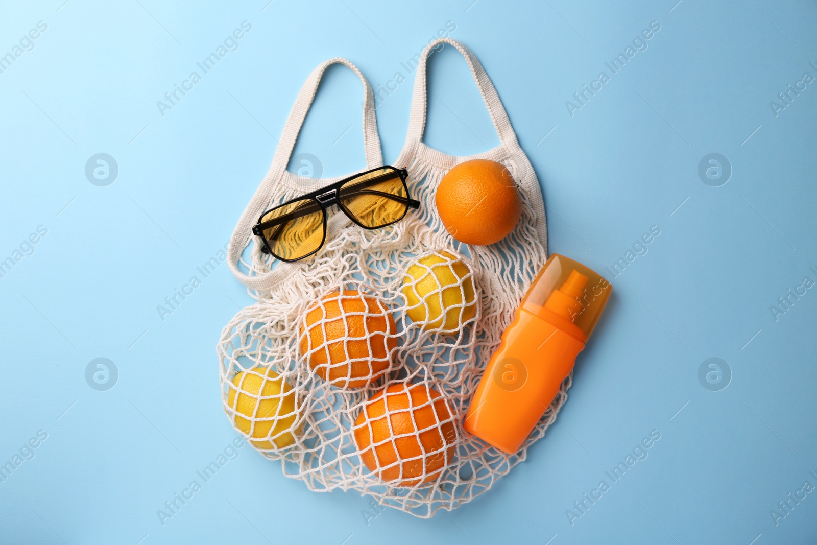 Photo of String bag with sunglasses, fruits and sunscreen on light blue background, flat lay