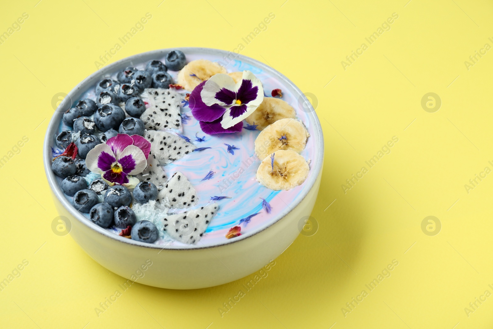 Photo of Delicious smoothie bowl with fresh fruits, blueberries and flowers on yellow background, closeup. Space for text