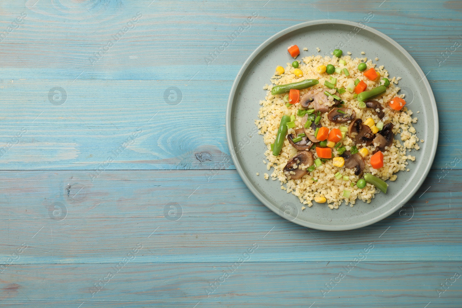 Photo of Delicious bulgur with vegetables and mushrooms on light blue wooden table, top view. Space for text