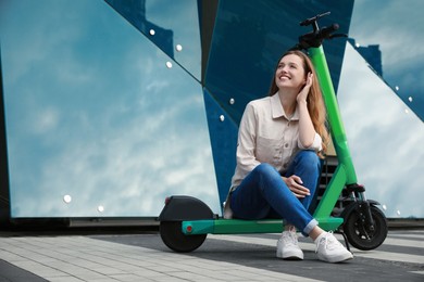 Happy woman sitting on modern electric kick scooter on city street, space for text