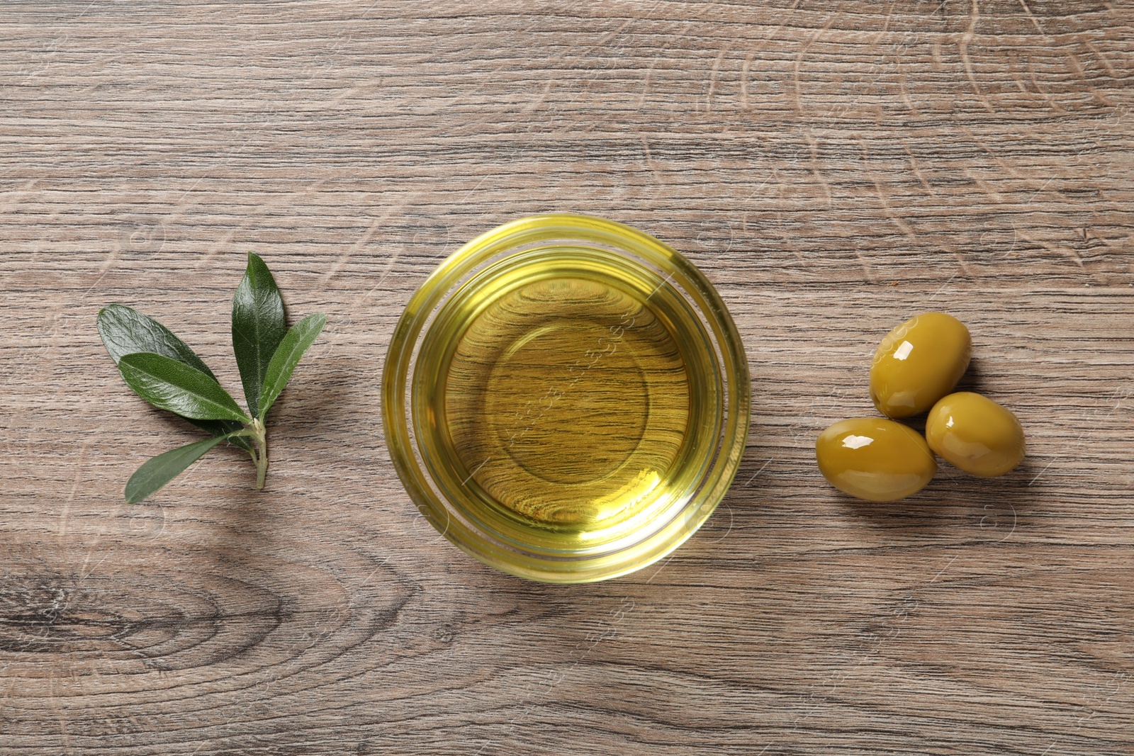 Photo of Glass bowl of oil, ripe olives and green leaves on wooden table, flat lay