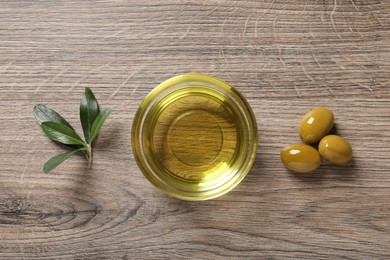 Photo of Glass bowl of oil, ripe olives and green leaves on wooden table, flat lay