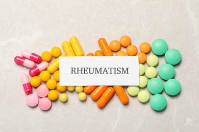 Photo of Paper with word Rheumatism and pills on light textured background, flat lay
