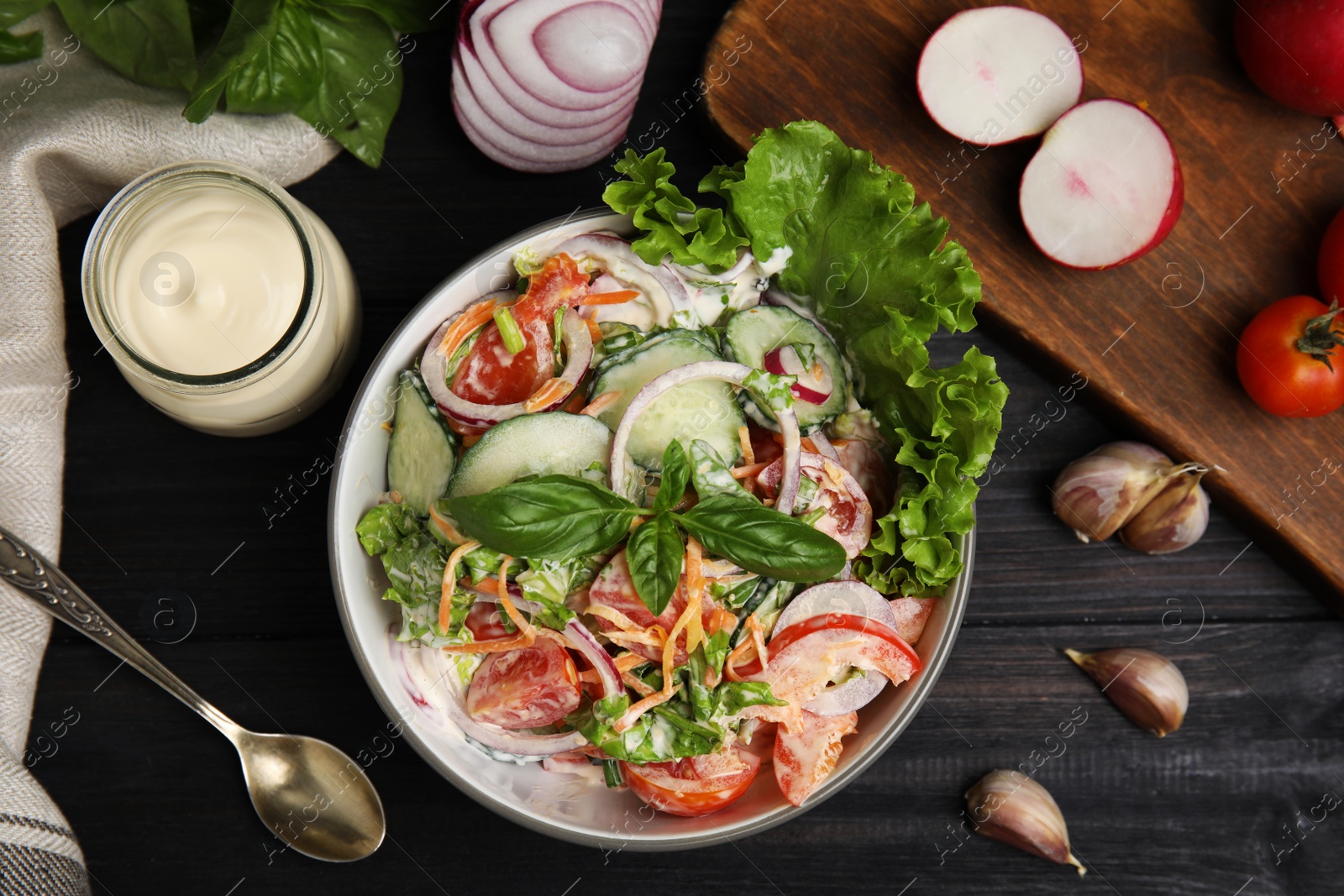 Photo of Bowl of delicious vegetable salad dressed with mayonnaise and ingredients on black wooden table, flat lay