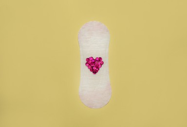 Photo of Sanitary pad with heart made of pink sequins on beige background, top view. Menstrual cycle
