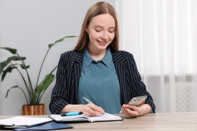 Photo of Woman taking notes while using smartphone at wooden table in office