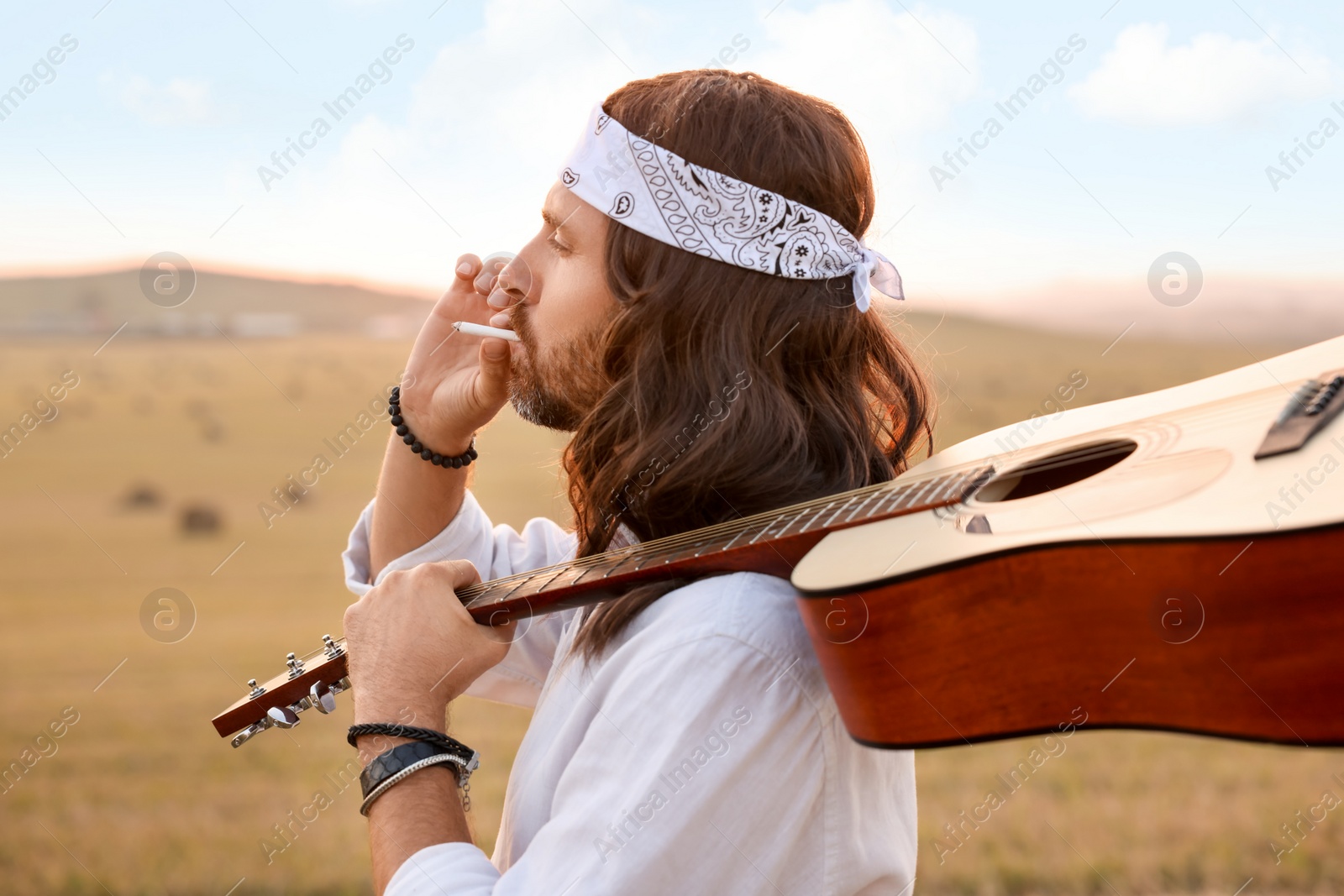 Photo of Stylish hippie man with guitar smoking joint in field