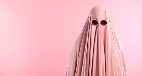 Photo of Glamorous ghost. Woman in sheet with sunglasses on pink background, space for text