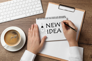 Image of Woman writing words NEW IDEA in notebook at wooden table, top view