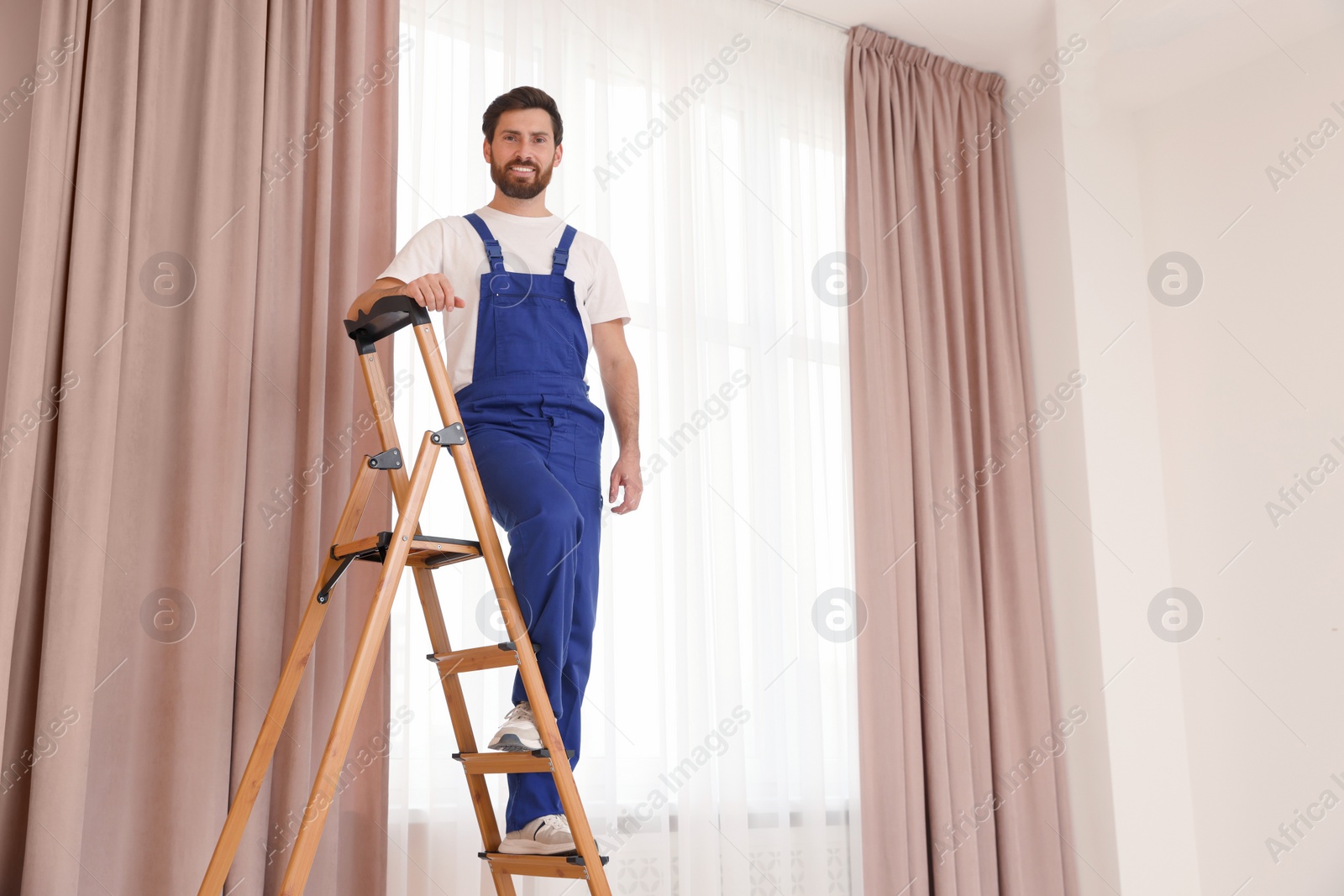 Photo of Worker in uniform standing on wooden folding ladder near window curtains indoors