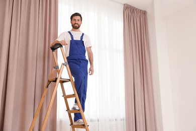 Worker in uniform standing on wooden folding ladder near window curtains indoors