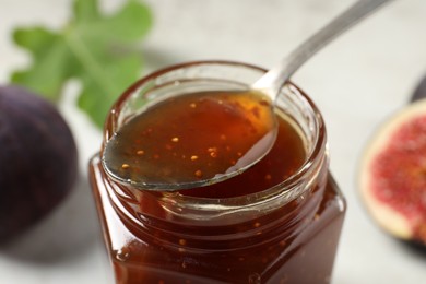 Photo of Glass jar of tasty fig jam with spoon against blurred background, closeup