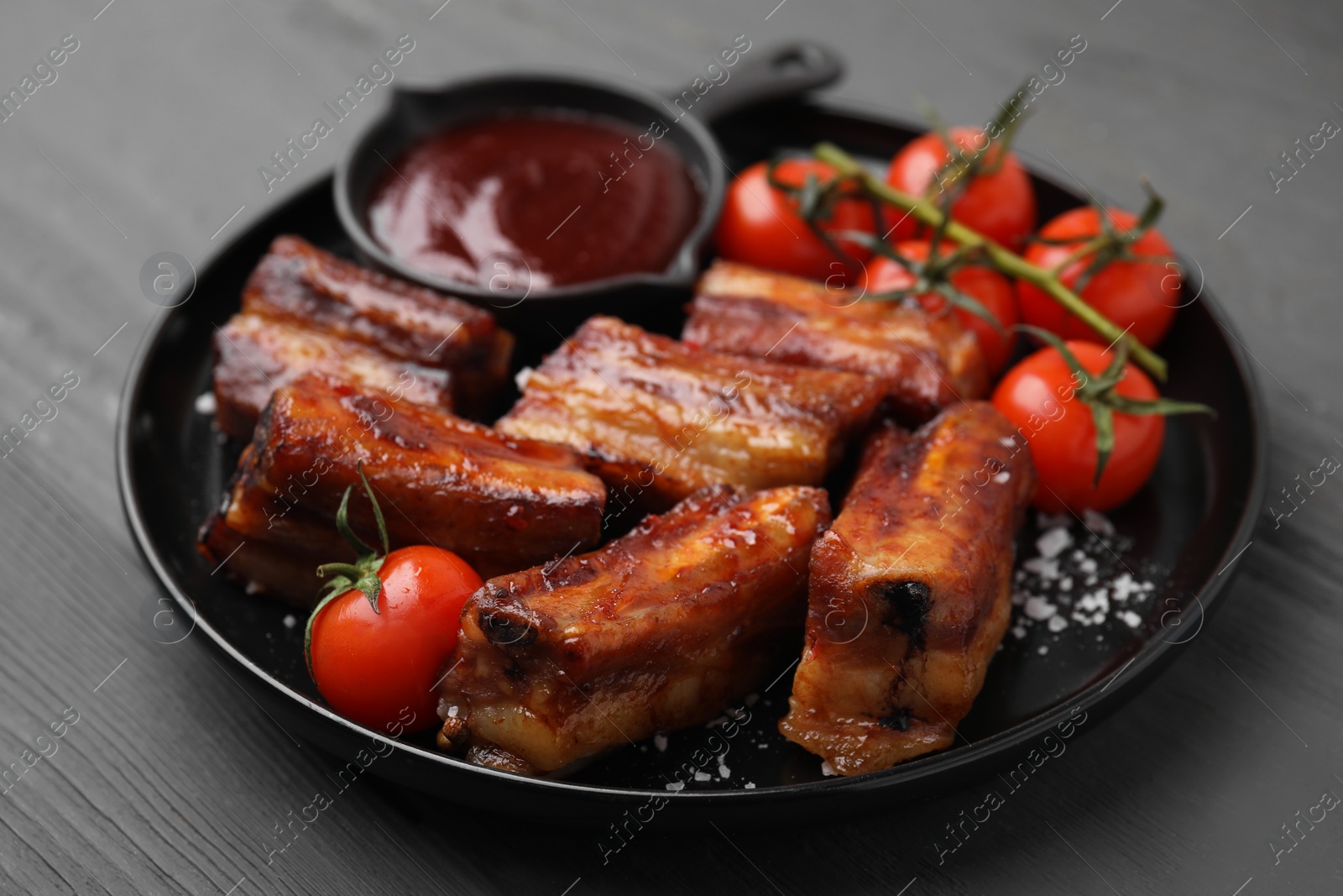 Photo of Tasty roasted pork ribs served with sauce and tomatoes on grey wooden table, closeup