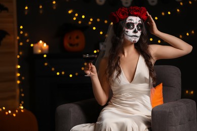 Photo of Young woman in scary bride costume with sugar skull makeup and glass of wine against blurred lights indoors, space for text. Halloween celebration