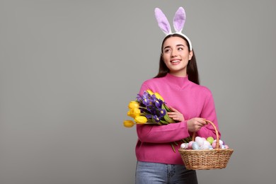 Happy woman in bunny ears headband holding wicker basket with painted Easter eggs and bouquet of flowers on grey background. Space for text