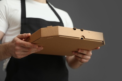 Waiter in apron with pizza box on grey background, closeup. Food delivery service