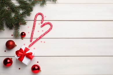 Tasty candy canes, Christmas balls, fir branches and gift box on white wooden table, flat lay. Space for text