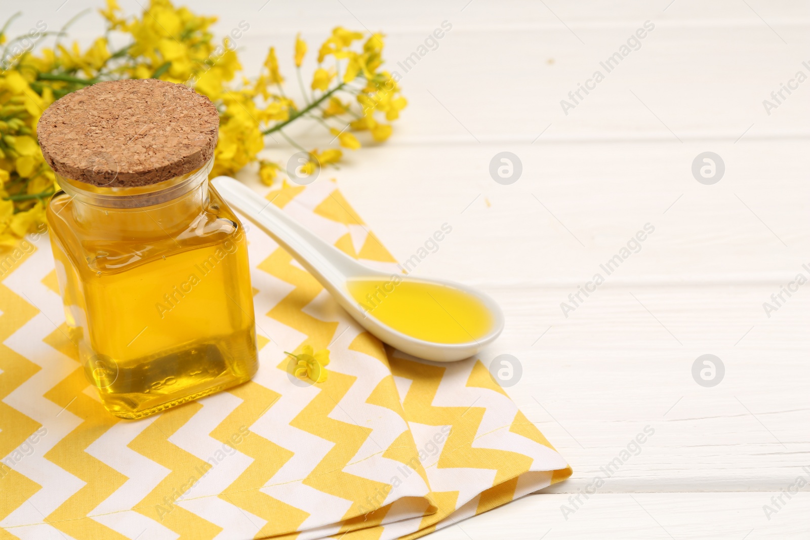 Photo of Rapeseed oil in glass bottle, gravy boat and beautiful yellow flowers on white wooden table, space for text
