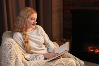 Photo of Beautiful young woman reading book near fireplace in room