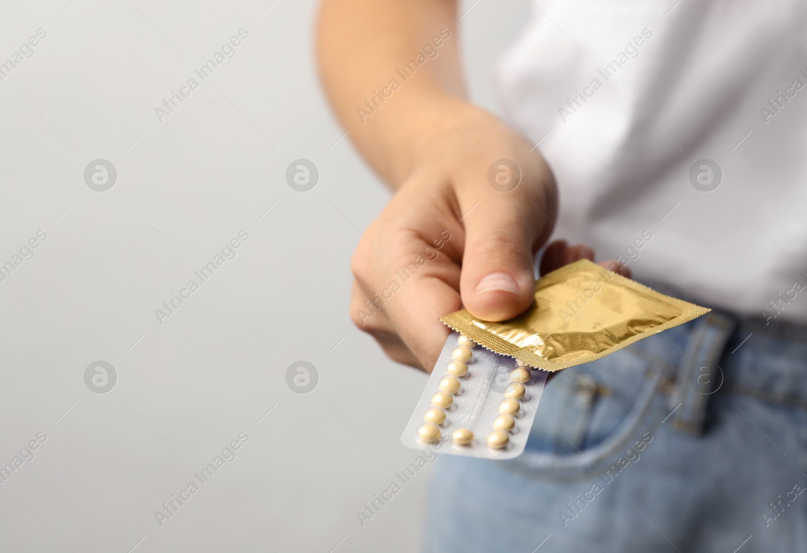 Photo of Young woman holding birth control pills and condom on light grey background, closeup with space for text. Safe sex concept