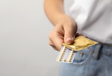 Young woman holding birth control pills and condom on light grey background, closeup with space for text. Safe sex concept