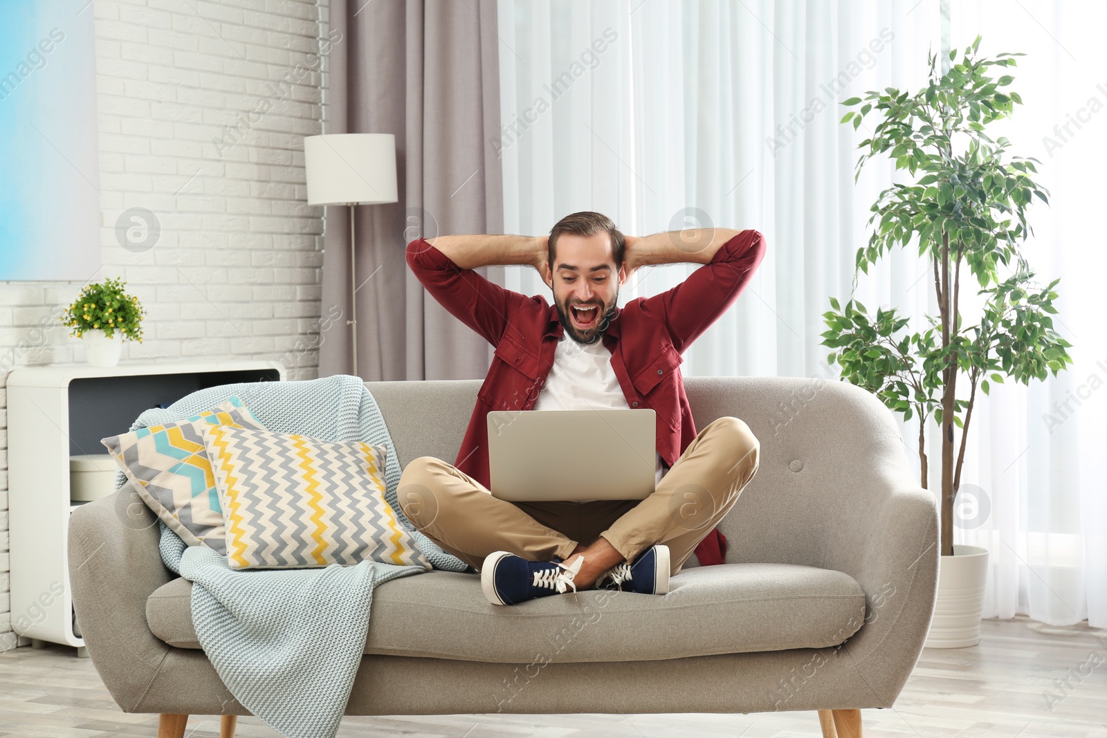 Photo of Emotional young man with laptop celebrating victory on sofa at home