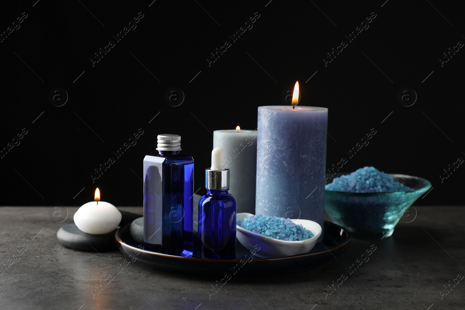 Photo of Spa composition. Cosmetic products, burning candles and sea salt on gray table against black background