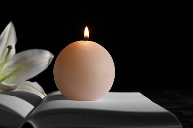 Burning candle and white lily on book in darkness, space for text. Funeral symbol