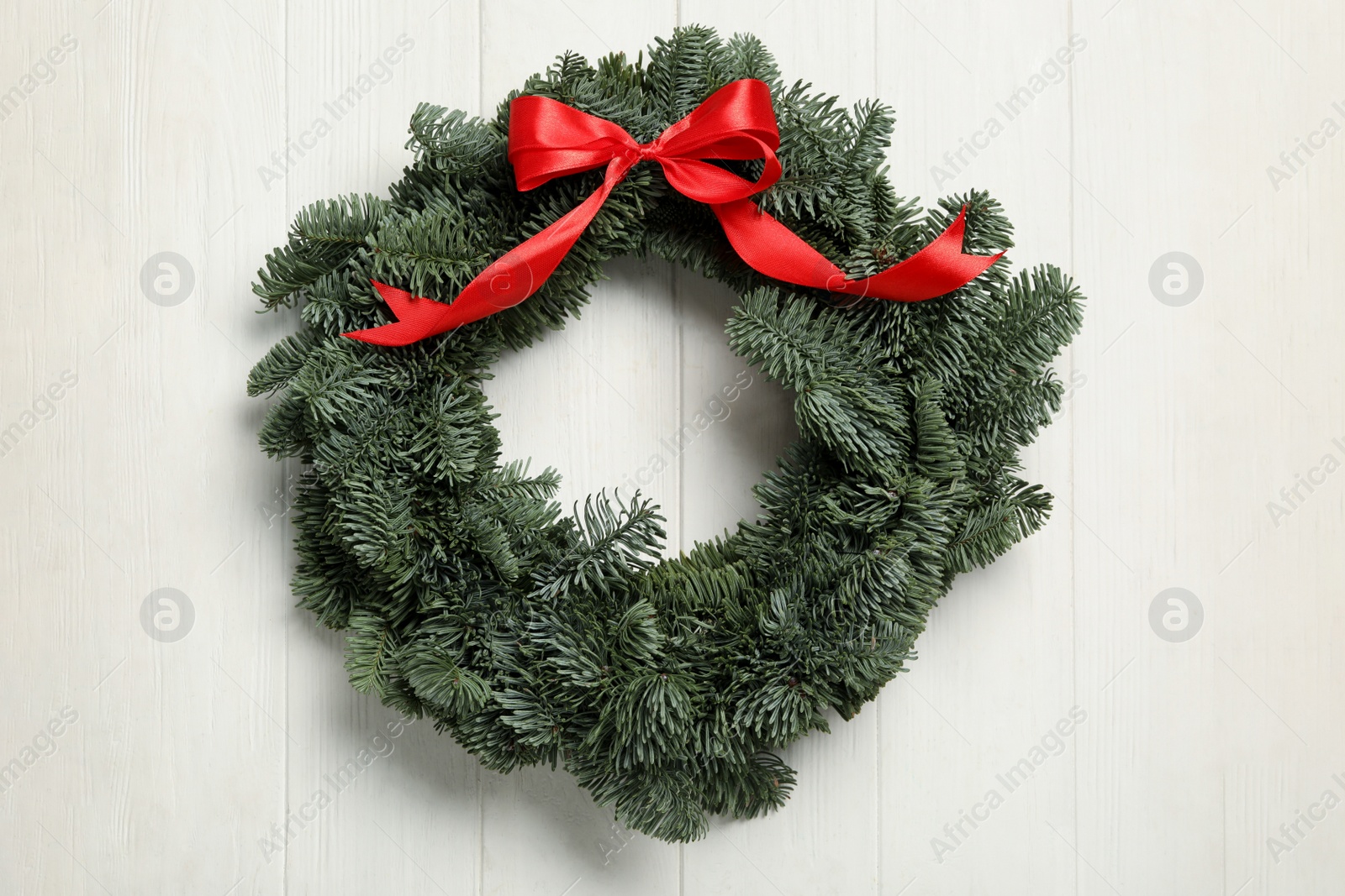Photo of Christmas wreath made of fir tree branches with red ribbon on white wooden background