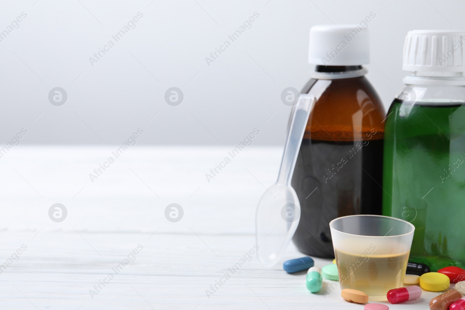 Photo of Bottles of syrup, measuring cup, dosing spoon and pills on white table against light grey background, space for text. Cold medicine