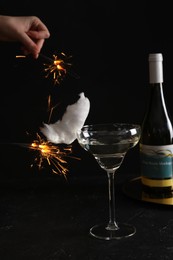 Photo of Cocktail with tasty cotton candy and bottle of alcohol drink on dark textured table. Woman holding sparkler against black background, closeup