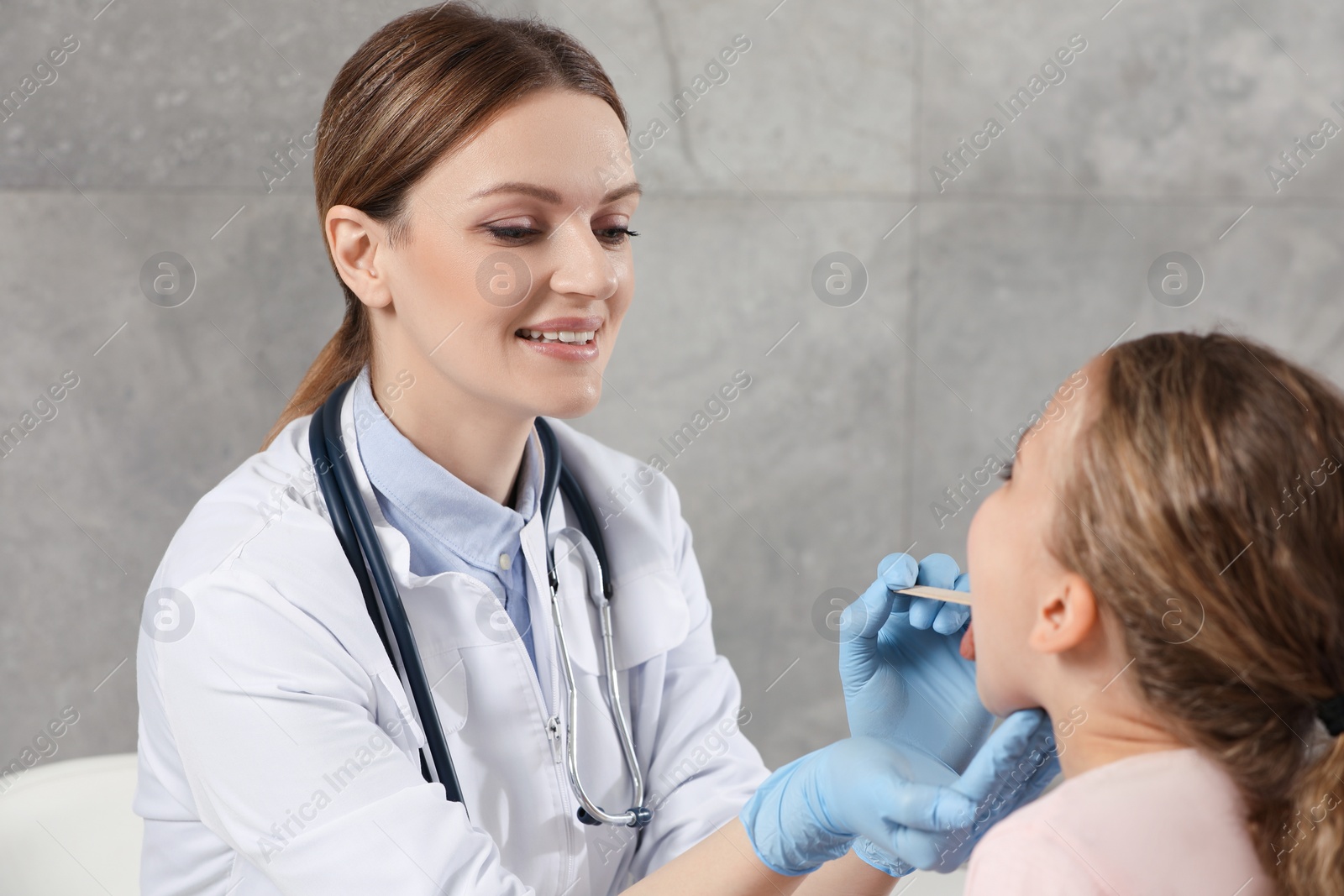 Photo of Smiling doctor examining girl`s oral cavity with tongue depressor indoors