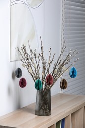 Photo of Beautiful pussy willow branches with paper eggs in vase on shelving unit at home. Easter decor
