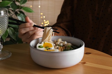 Woman eating delicious ramen with chopsticks at wooden table indoors, closeup. Noodle soup