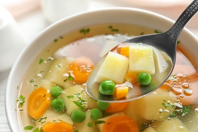 Photo of Spoon of fresh homemade vegetable soup over full bowl on table, closeup