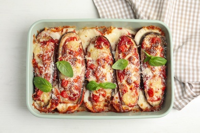 Baked eggplant with tomatoes, cheese and basil in dishware on white wooden table, top view