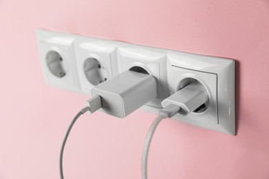Photo of Charger adapters plugged into power sockets on pink wall, closeup. Electrical supply