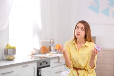 Thoughtful young woman choosing between orange and donut in kitchen, space for text. Healthy diet