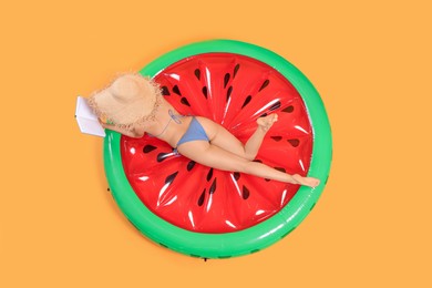 Sexy young woman with beautiful suntan and hat reading book on inflatable mattress against orange background, top view